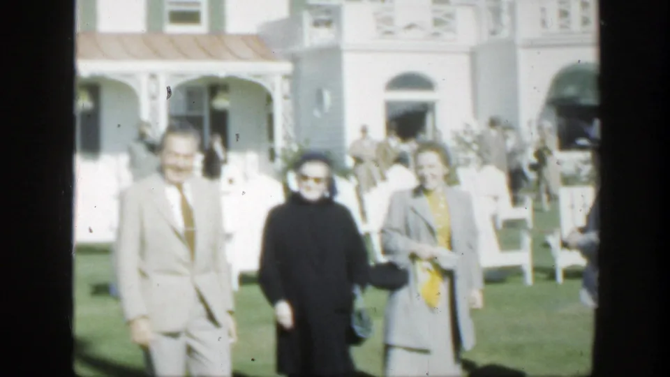 1950 Montreal frame of adults walking toward the camera on a sunny lawn outside a white house.