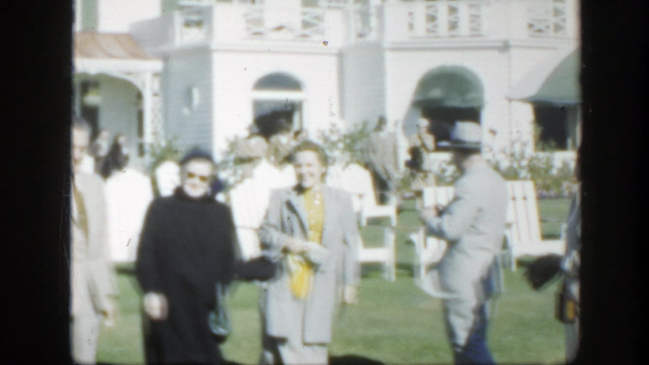 1950 Montreal home-movie frame of adults in coats and suits standing on a lawn before a white building with green awnings.