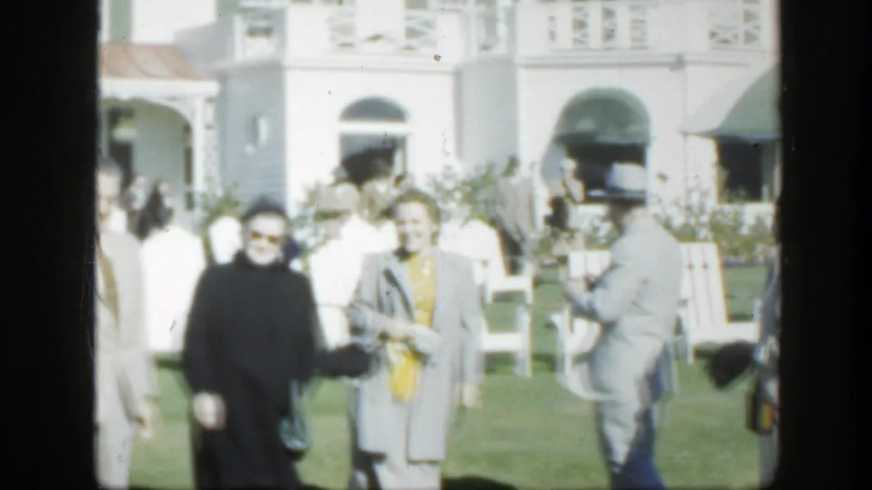 1950 Montreal home-movie frame of adults in coats and suits standing on a lawn before a white building with green awnings.