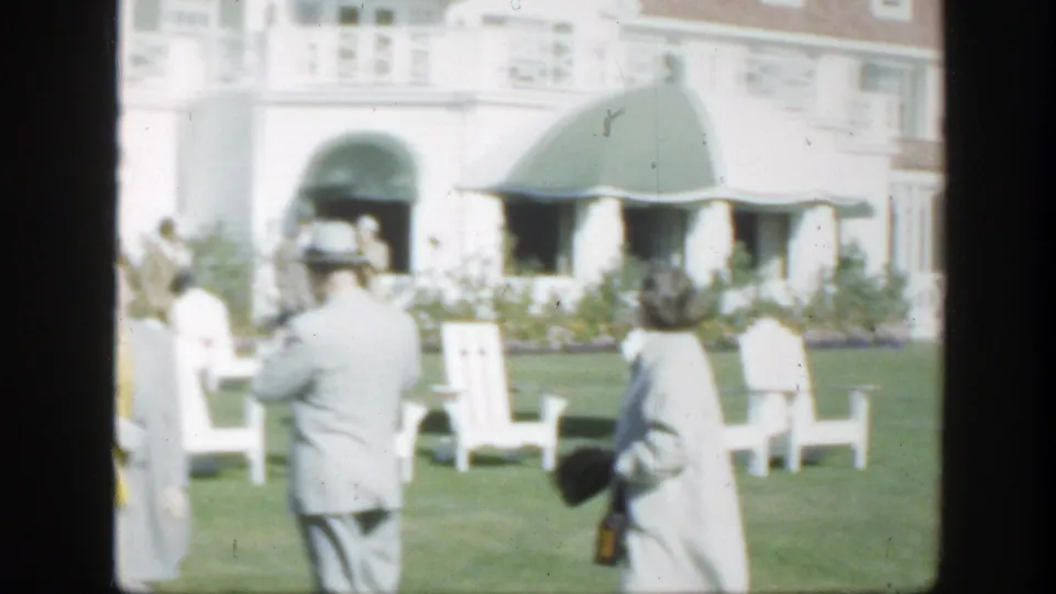 Adults stand and move across a sunny lawn in Montreal, with white chairs, green awnings, and a pale building facade behind them.