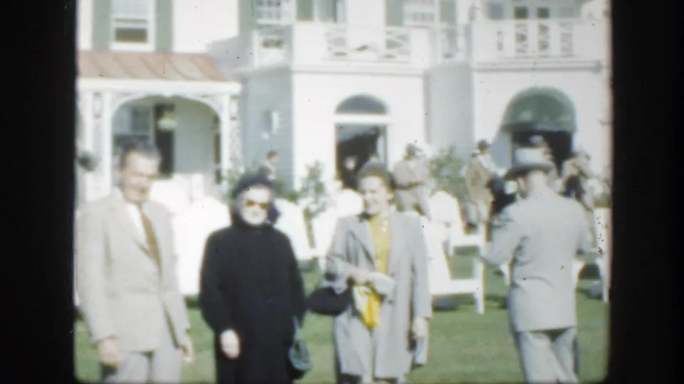1950 Montreal home-movie frame showing adults in mid-century dress facing the camera on a sunny lawn outside a white building.