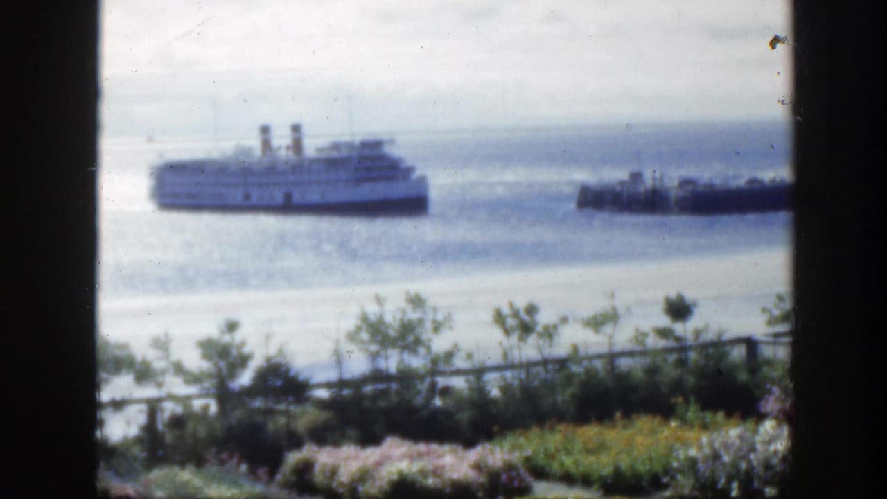 1950 Montreal 8mm frame showing a twin-funnel passenger ship offshore, a smaller boat, calm water, garden shrubs, railing, and dark foreground framing.