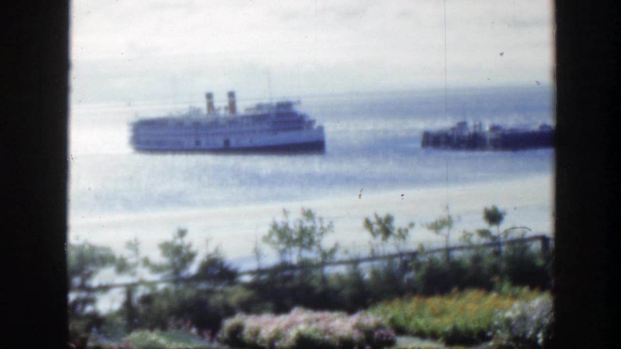 The passenger ship remains offshore in this 1950 Montreal home-movie frame, with a smaller boat to the right and garden plantings below.