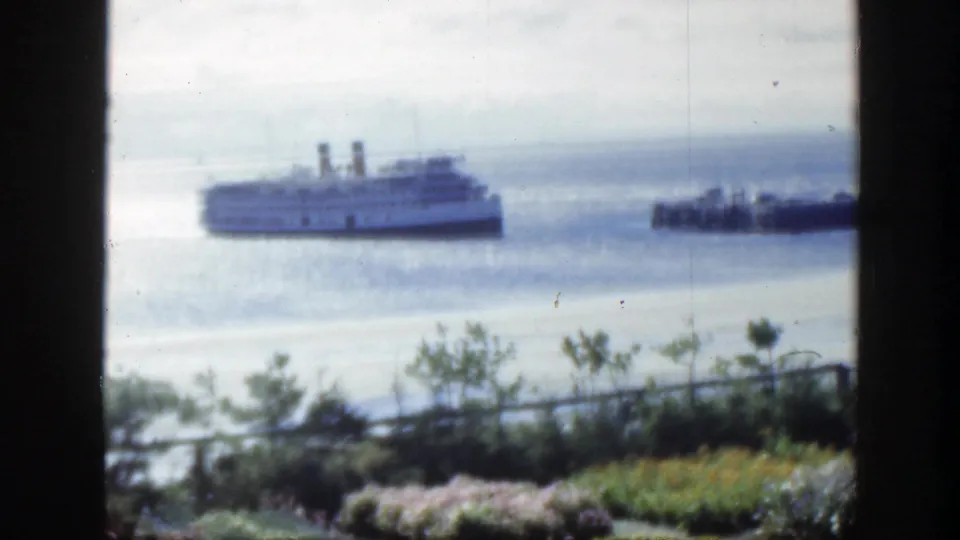 The passenger ship remains offshore in this 1950 Montreal home-movie frame, with a smaller boat to the right and garden plantings below.