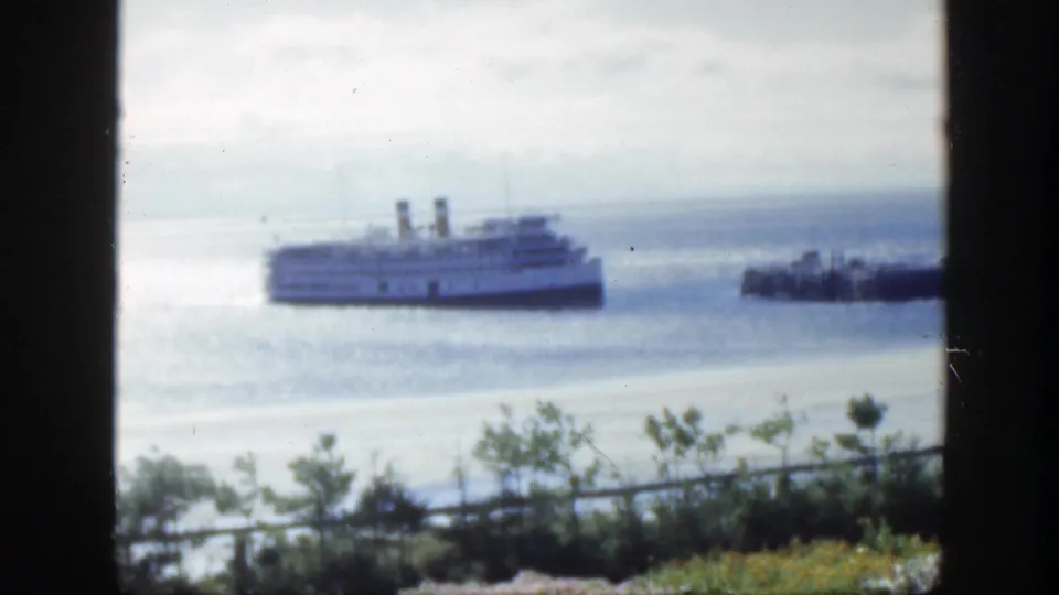 Late frame of the 1950 Montreal clip with the passenger ship, smaller boat, smooth water, overcast sky, foreground greenery, and black frame edge.