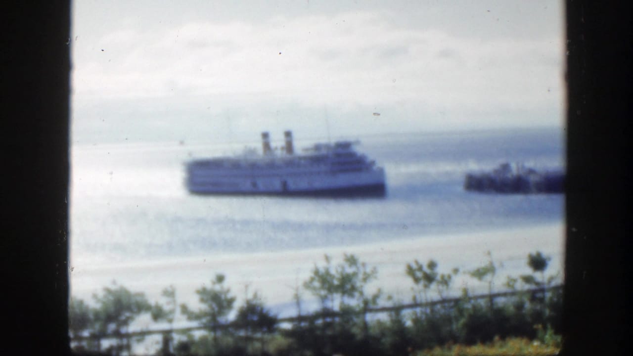 1950 home-movie view in Montreal, Canada, showing a large passenger ship offshore through dark vertical framing, with shrubs and shoreline in front.