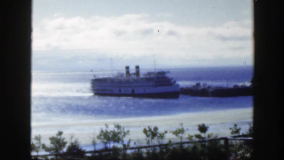 1950 Montreal home-movie still showing the passenger ship near shore through dark framing, with rippled water, cloudy sky, and shrubs below.