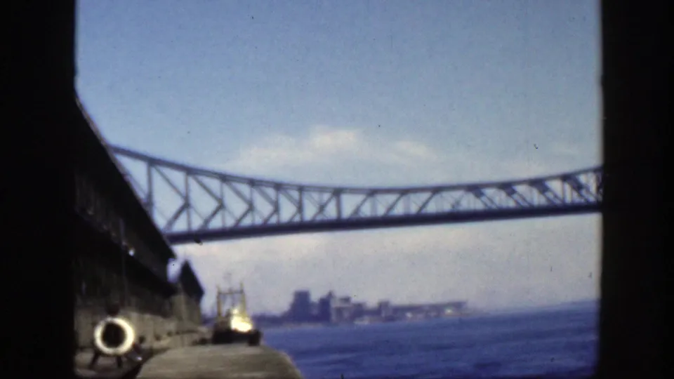 1950 Montreal-area 8mm view of a steel truss bridge over a broad waterway, with a small boat and distant city buildings below.