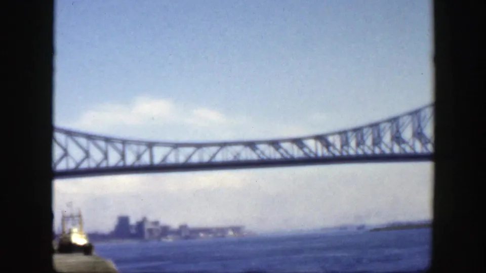 1950 Montreal-area frame showing a steel truss bridge stretching over water, with a small vessel and distant waterfront skyline beneath it.