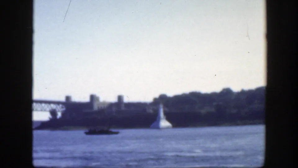 1950 Montreal 8mm frame with the bridge receding left, a small boat on the river, waterfront buildings, trees, and a white lighthouse-like structure.