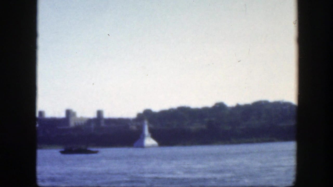 1950 Montreal waterfront view from a moving vessel, with a hazy river, distant shoreline, white conical shore structure, and small boat.