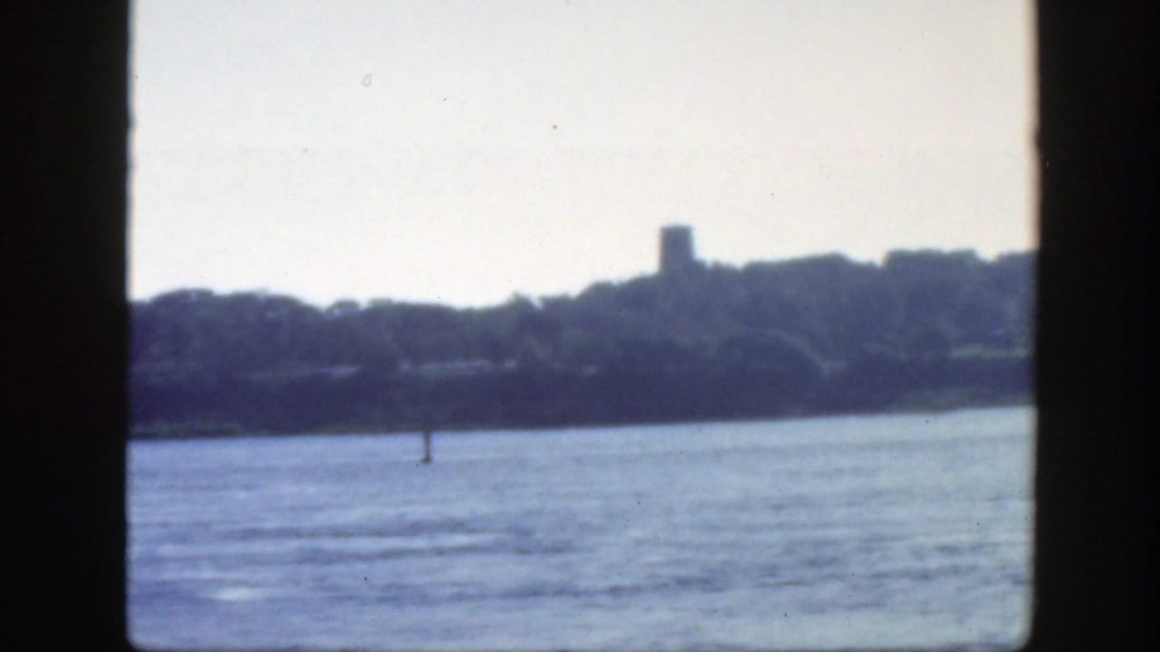 1950 Montreal-area 8mm frame showing calm glistening water, a wooded shoreline, and a distant tower-like structure beyond the shore.