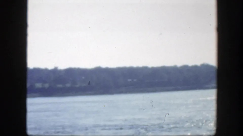 1950 Montreal home-movie view across blue water toward a low wooded shoreline, with gate edges and grain visible.