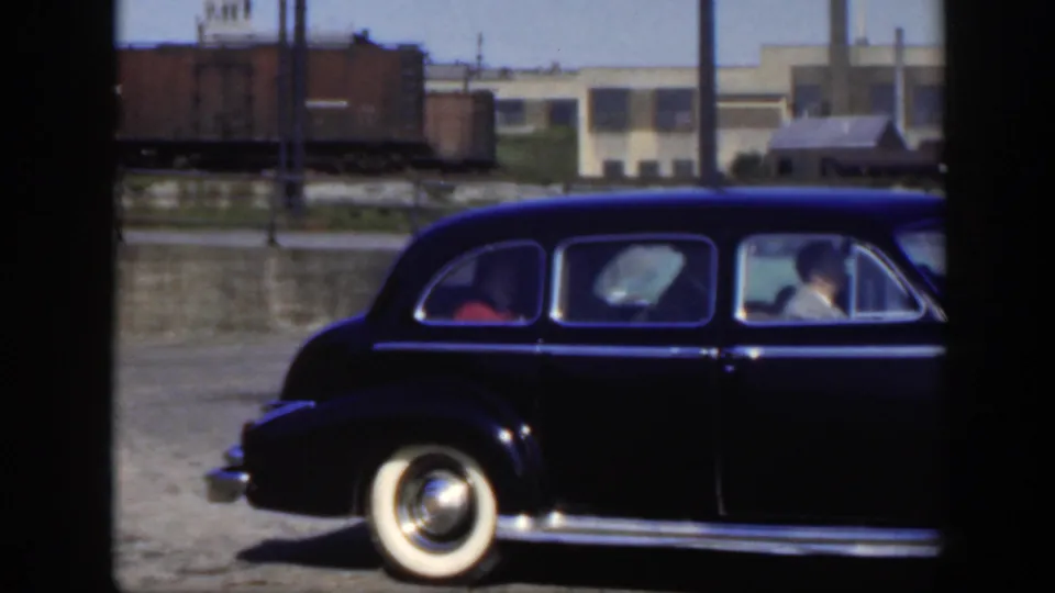 1950 Montreal frame of a black mid-century sedan near rail cars and industrial buildings, with occupants visible through the windows.