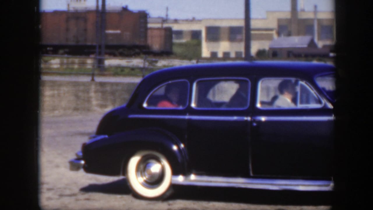 Black sedan with whitewall tires passing in front of a rail yard.