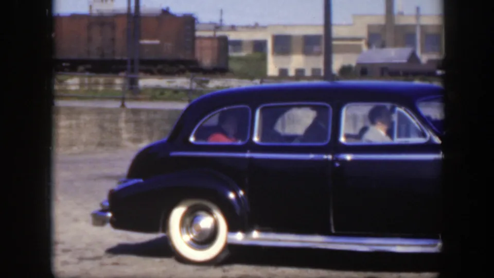 1950 Montreal home-movie frame of a black sedan moving past freight cars and industrial buildings.