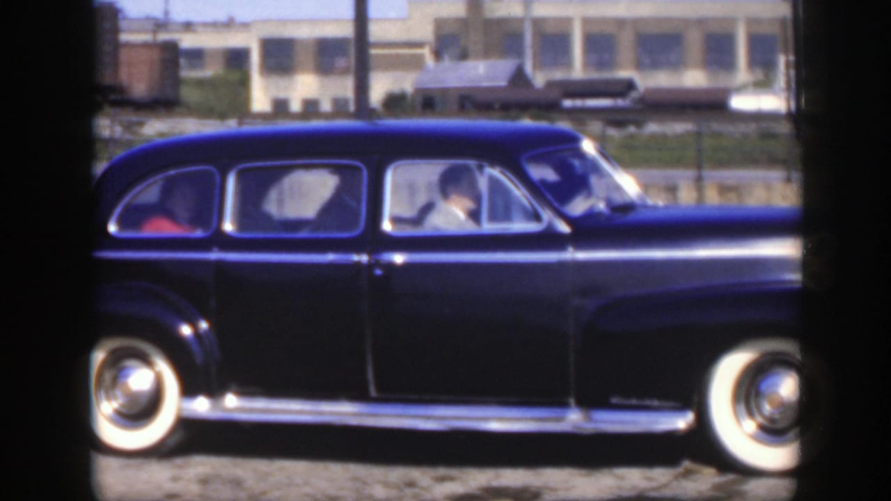 Side view of a black sedan with a driver visible near freight cars.