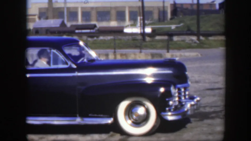 Close side view of the sedan's front section, with chrome grille, whitewall tire, and rail-yard buildings behind.