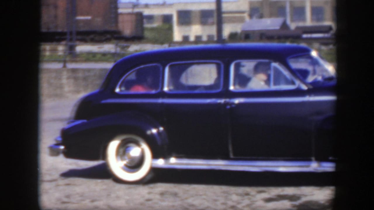 Black sedan with whitewall tires passes on a road with driver and passenger visible.
