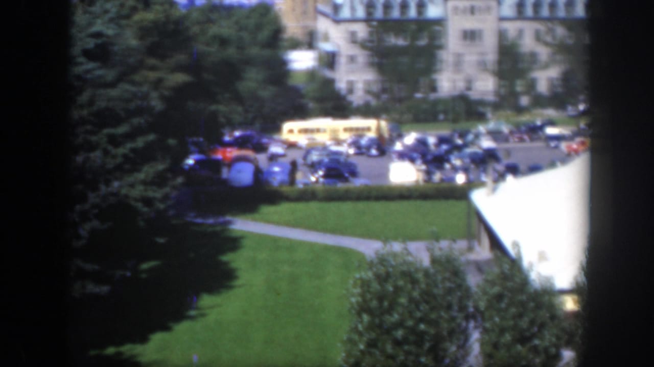 Elevated view of parked cars, a yellow bus, lawns, trees, and large buildings.