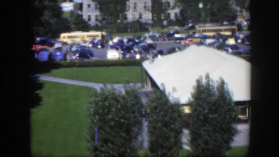 1950 Montreal home-movie frame of a parking area with cars and a yellow bus beyond lawns, trees, and a light roof.