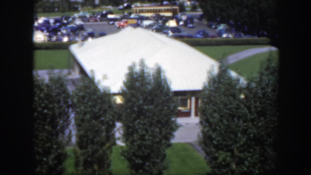 White-roofed building behind tall trees with parked cars in the distance.