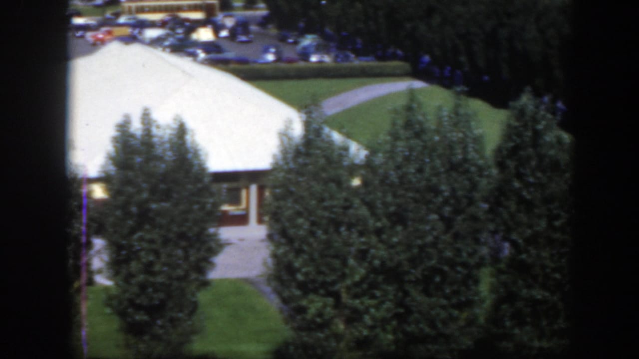 White-roofed building, curved paths, trees, lawn, and distant parked cars.