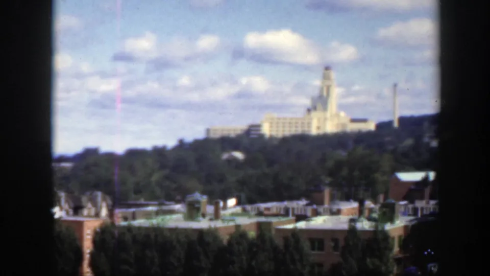 1950 Montreal home-movie frame showing a pale hilltop building beyond trees and rooftops under a blue, clouded sky.