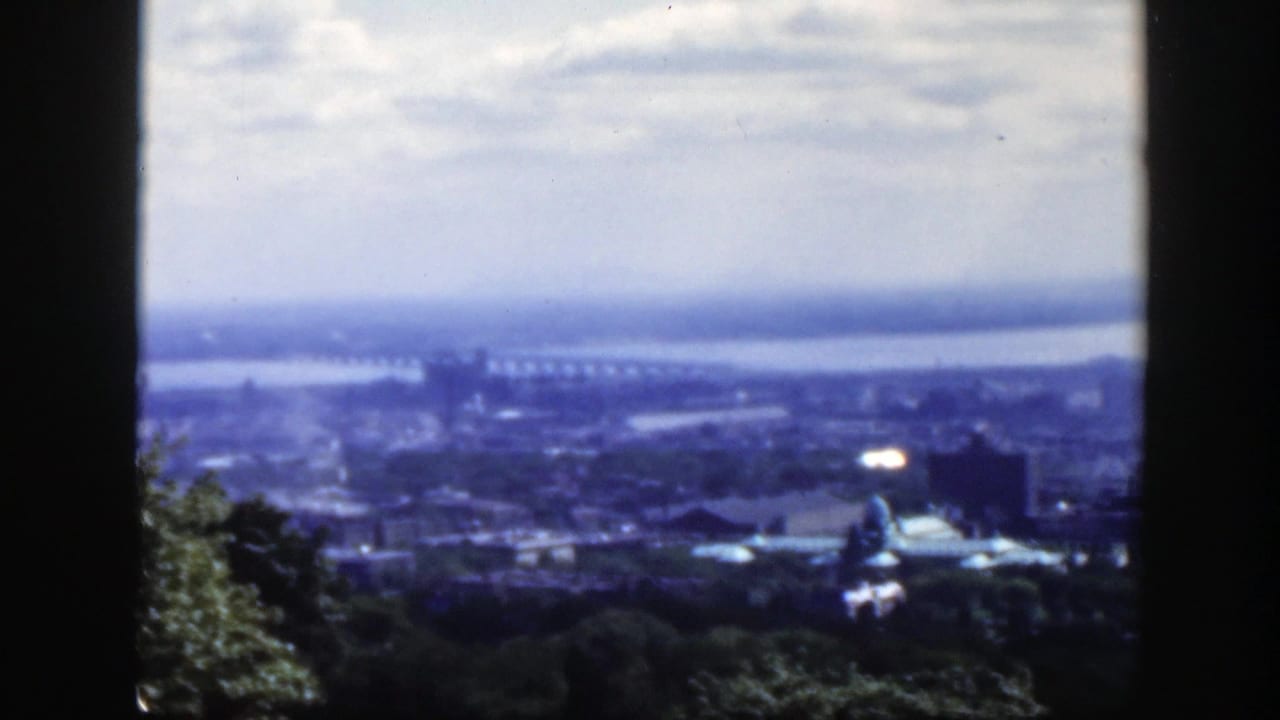 1950 Montreal home-movie view from above showing a hazy skyline, broad waterway, long bridge, domed structure, cloudy sky, and dark framing edges.