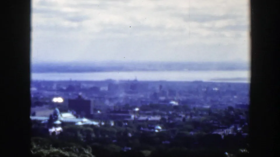 Distant 1950 Montreal skyline under cloudy sky, with water and bridge in the background, a domed building in the lower city, and dark vertical edges framing the view.