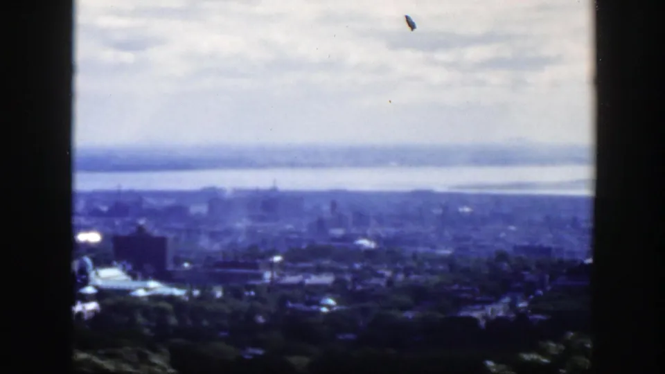 1950 Montreal city panorama seen from an elevated vantage, with dark mullions, foreground trees, low buildings, distant skyline, and river haze.