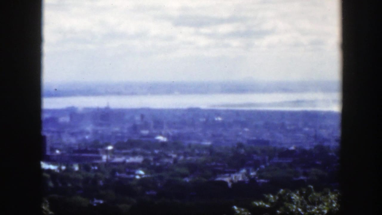Elevated 1950 Montreal view through dark window framing, showing a hazy skyline, residential foreground, trees, river, and overcast sky.