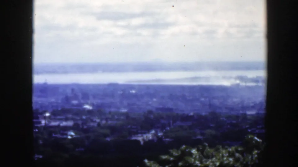 1950 Montreal cityscape from a high viewpoint, framed by a dark mullion, with trees, buildings, river, cloudy sky, and faint smoke or haze.