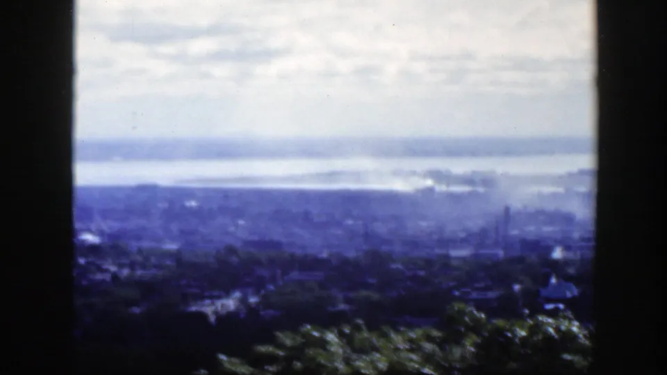 1950s Montreal home-movie frame showing a hazy city and distant water from an elevated viewpoint behind dark vertical bars.