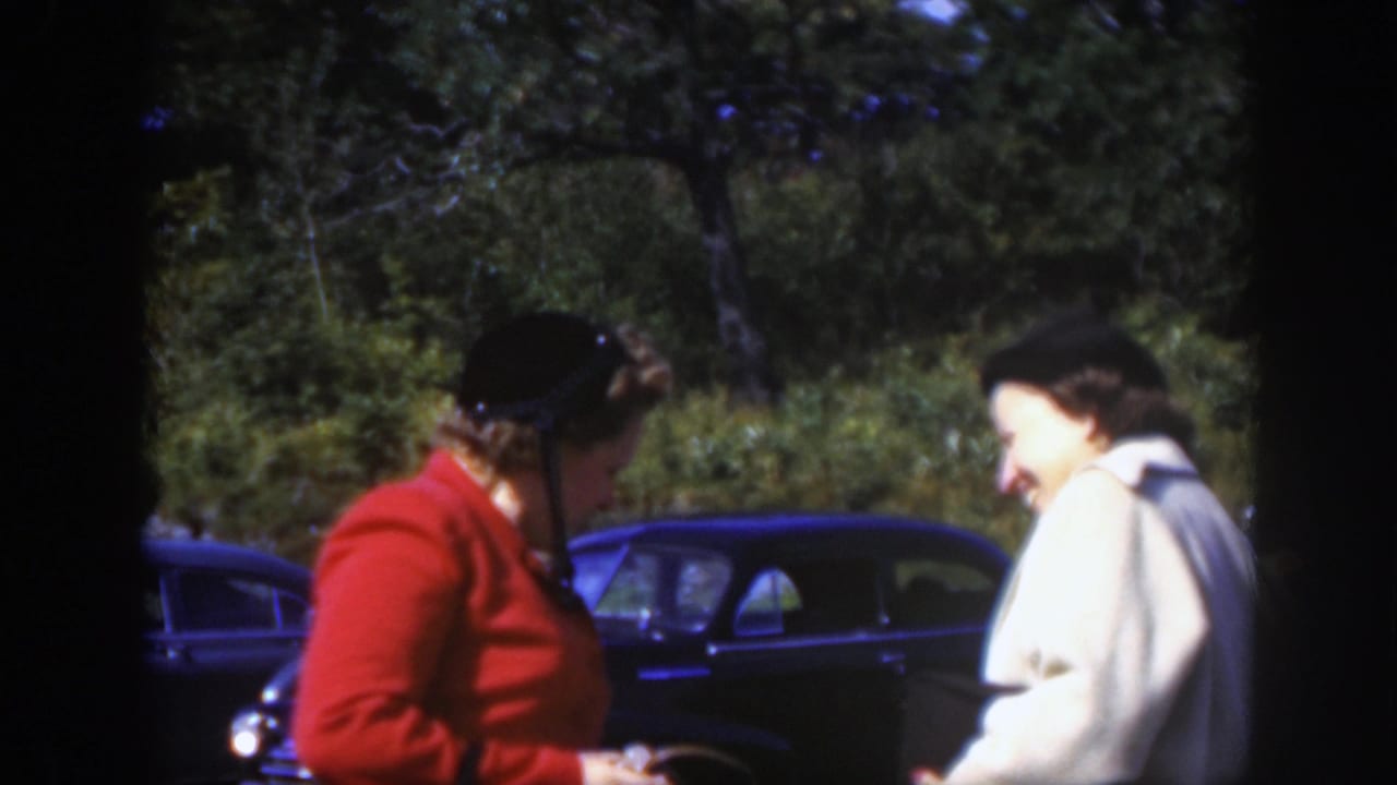Two women in hats talk outdoors in Montreal, one in a red jacket and one in a light coat, with dark parked cars behind them.