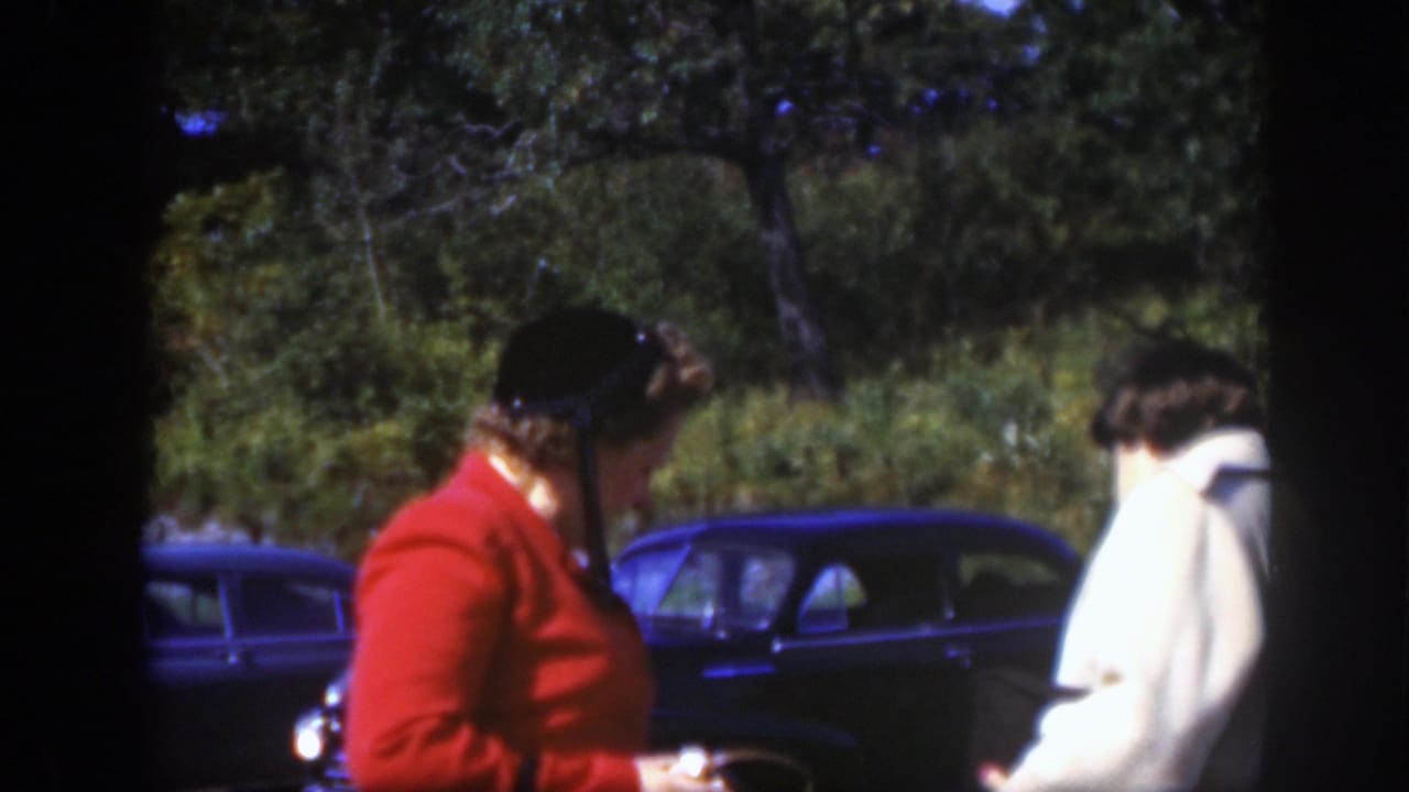 In this 1950 Montreal frame, a woman in a red jacket looks toward a woman in a light coat near dark cars and trees.