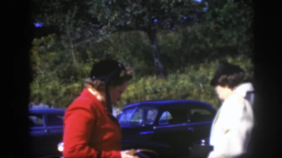 Two hat-wearing women in Montreal stand close together, looking down as they talk beside a dark mid-century car.