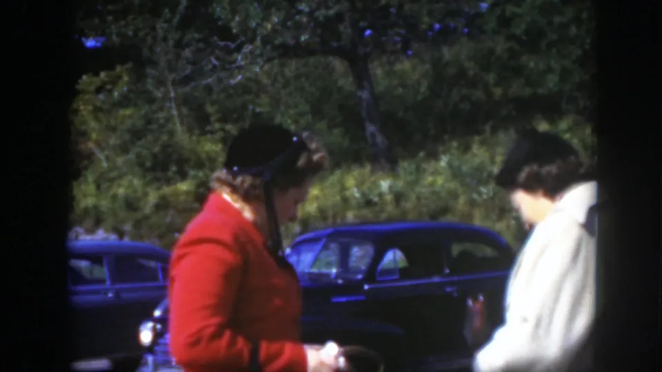 A woman in a red jacket stands at left while a woman in a light coat turns away, with a dark parked car and trees in the background.