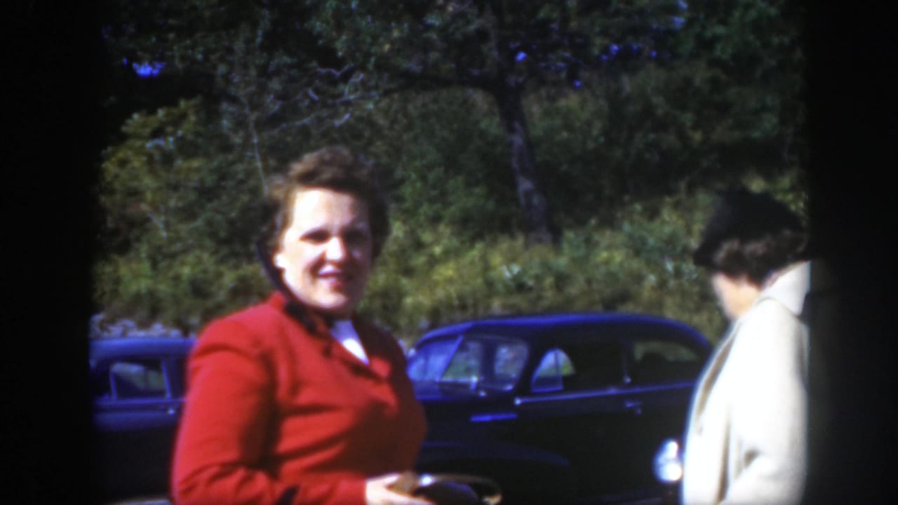 Woman in a red jacket stands near parked dark cars on a sunny tree-lined street.
