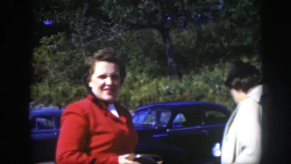 In supported 1950 Montreal home-movie footage, a woman in a red jacket stands near dark parked cars with trees behind her.