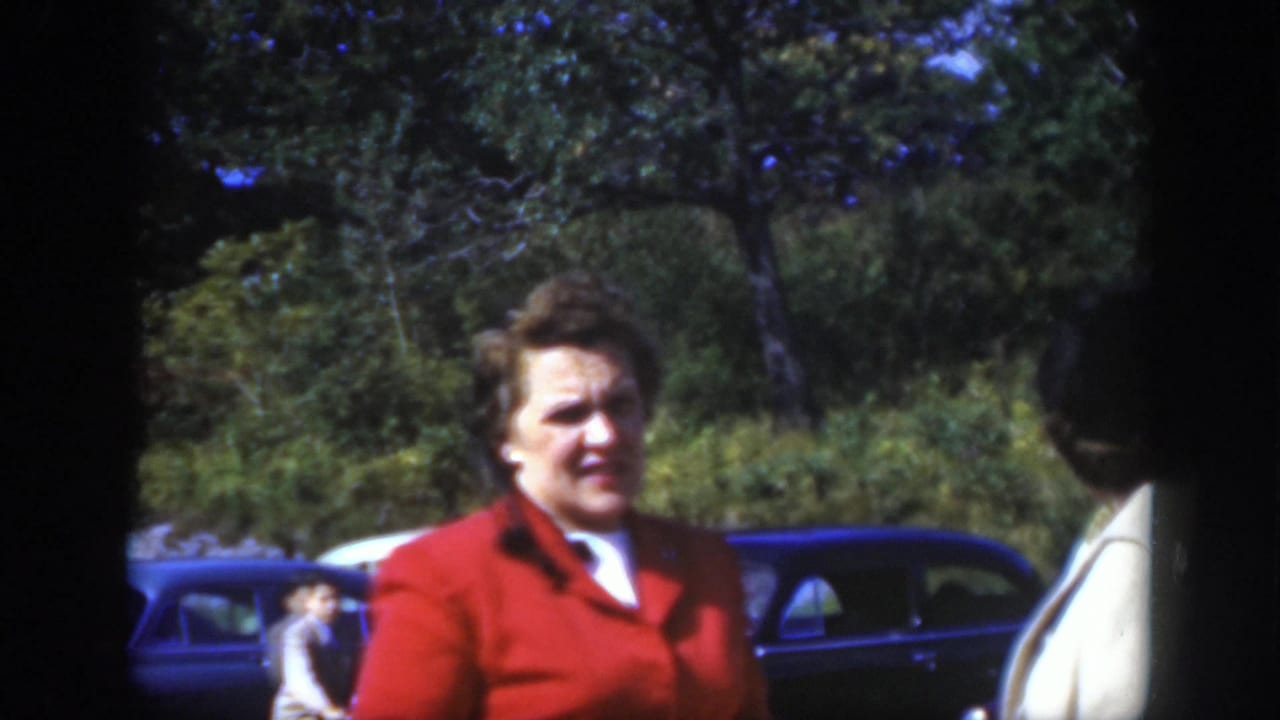 The woman in red faces the camera in the 1950 Montreal street scene as another adult stands near parked automobiles.