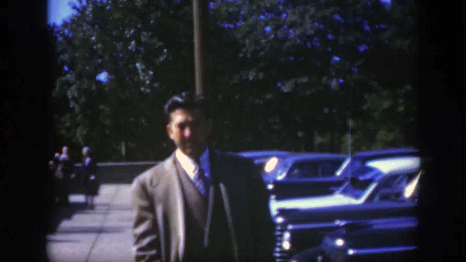 A man in a suit and light overcoat stands on a Montreal sidewalk near classic cars in the supplied 1950 home-movie footage.