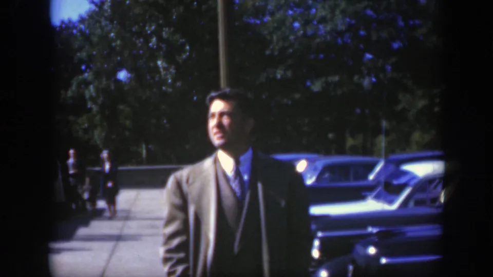 The suited man looks away from the camera beside parked cars, with trees and distant pedestrians in the 1950 Montreal street setting.