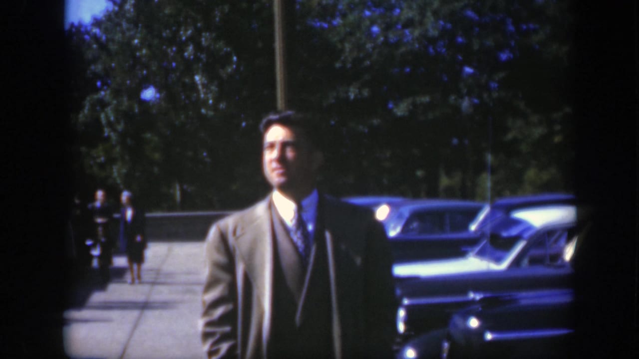 Suited man standing near parked cars with pedestrians in the background.