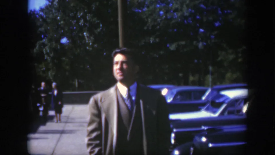 A suited adult man stands in a Montreal parking area circa 1950, with classic cars, trees, and distant pedestrians behind him.