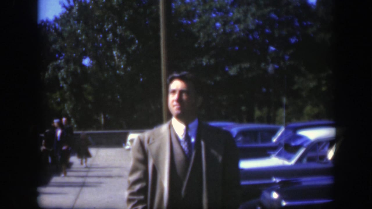 The suited man remains near the parked cars in Montreal, with a tree-lined background and distant people on the walkway.
