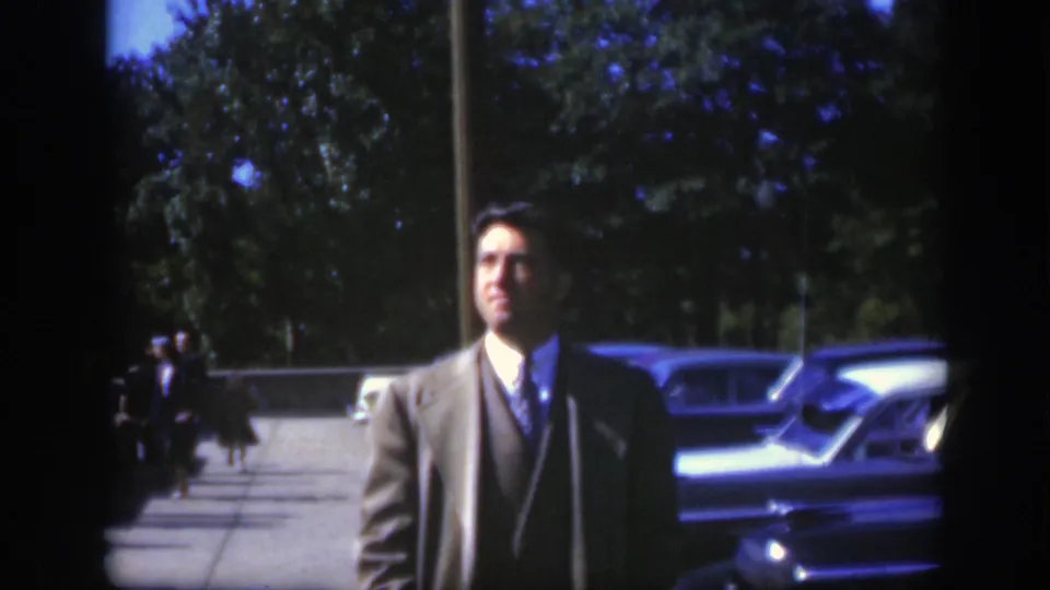 The suited man remains near the parked cars in Montreal, with a tree-lined background and distant people on the walkway.