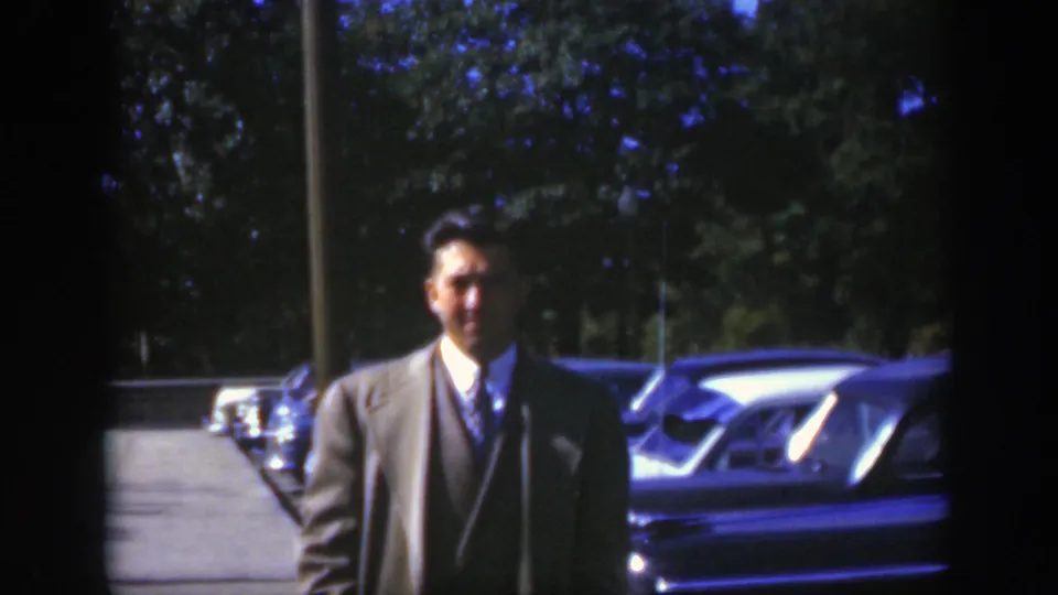 A well-dressed man faces near the camera in the parking area, with chrome-trim cars and trees visible behind him.