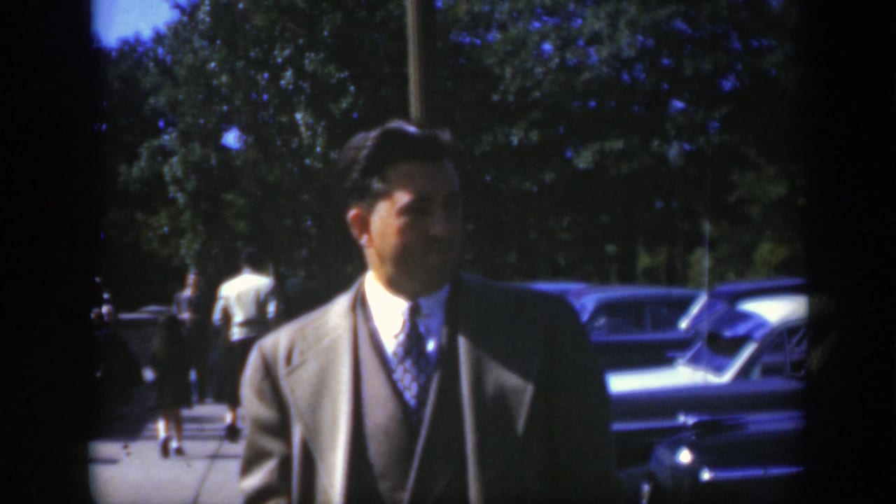 In Montreal circa 1950, a man in a light-colored suit and patterned tie stands outdoors near parked mid-century cars and trees.