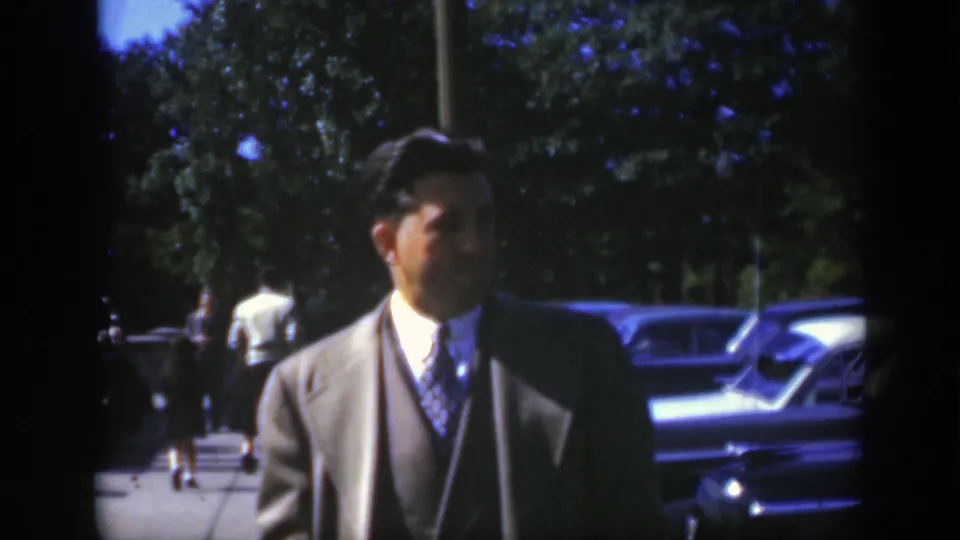 In Montreal circa 1950, a man in a light-colored suit and patterned tie stands outdoors near parked mid-century cars and trees.